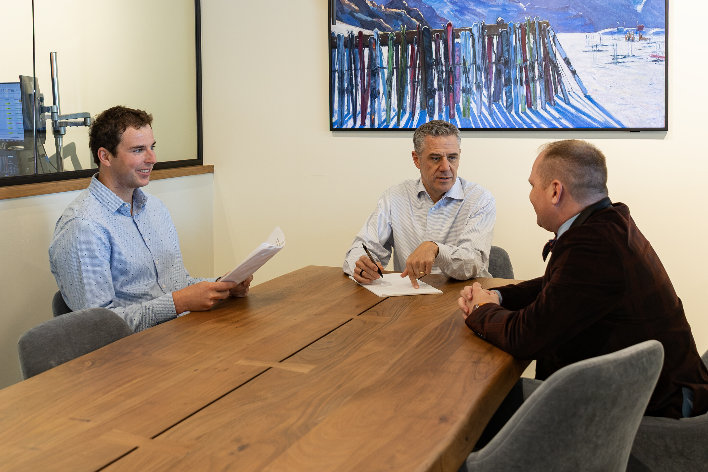 Three individuals are sitting at a table engaged in a discussion, with papers and a computer nearby.