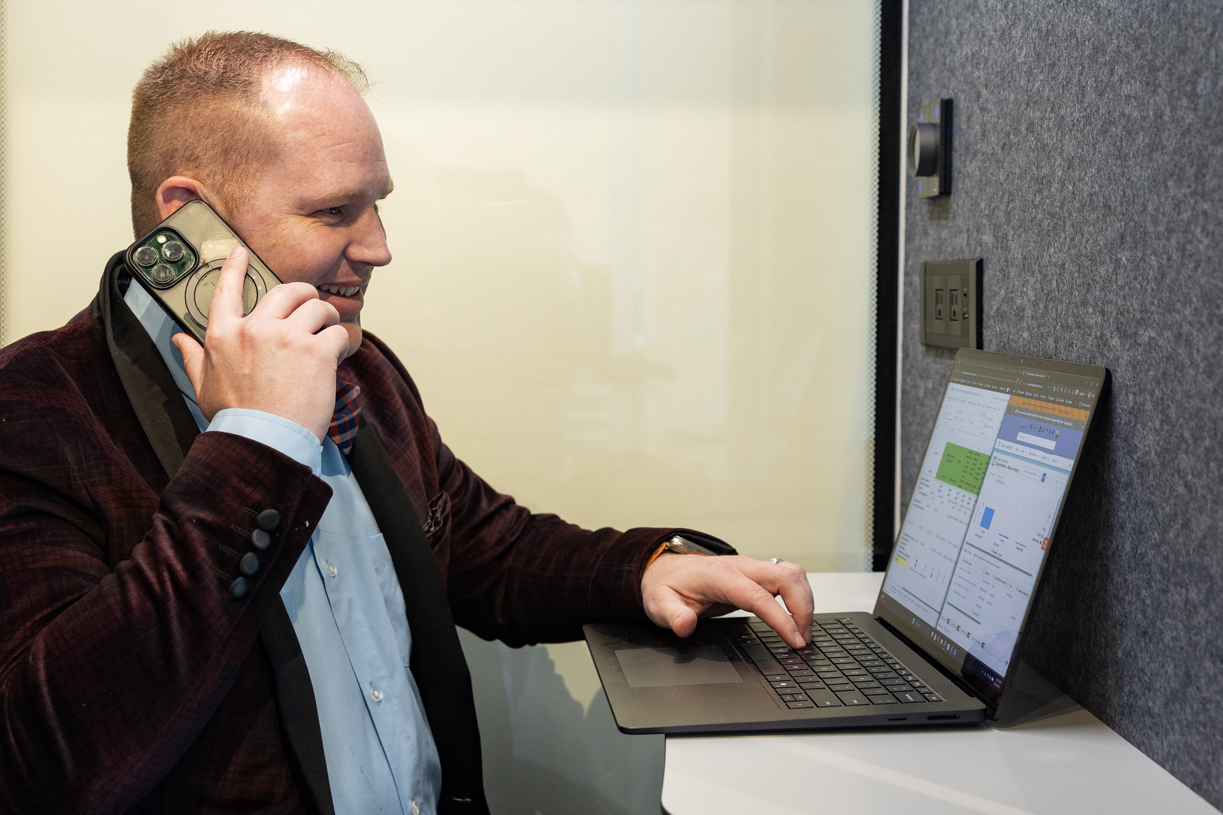 Man in a suit talking on the phone while typing on a laptop at a desk.