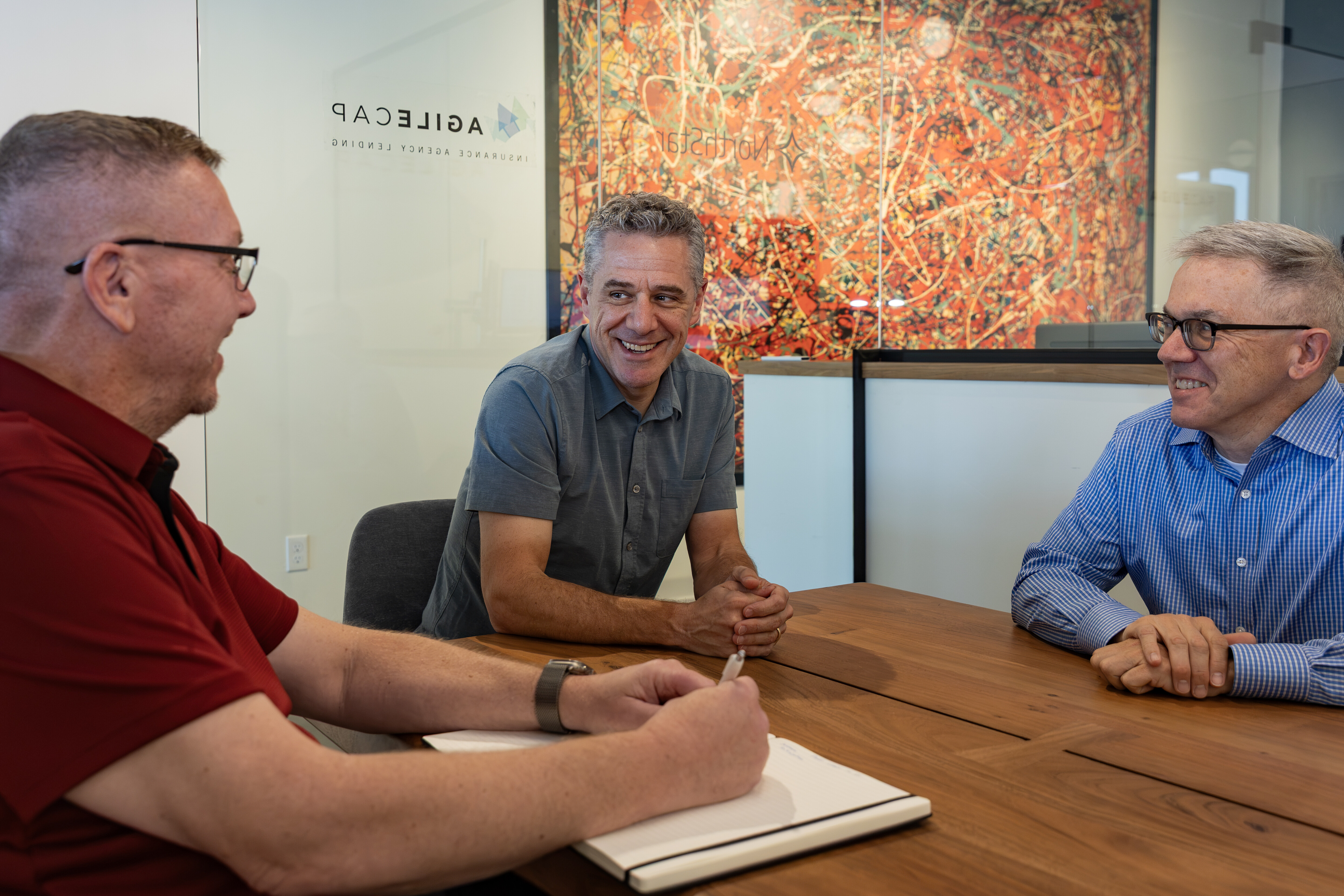 Three men engage in a discussion at a wooden table in an office setting.