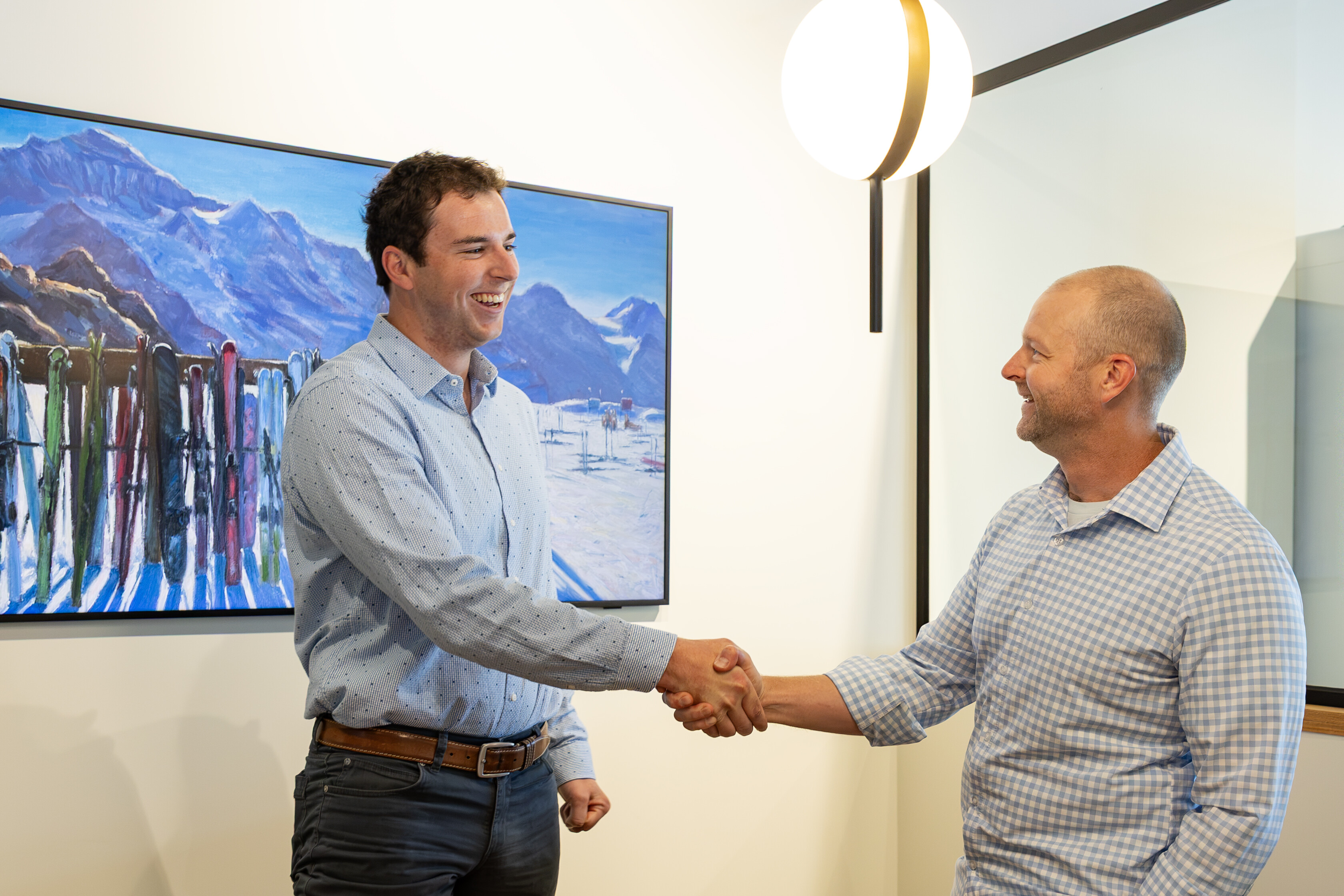 Two men shake hands and smile in a bright room with a ski-themed painting.