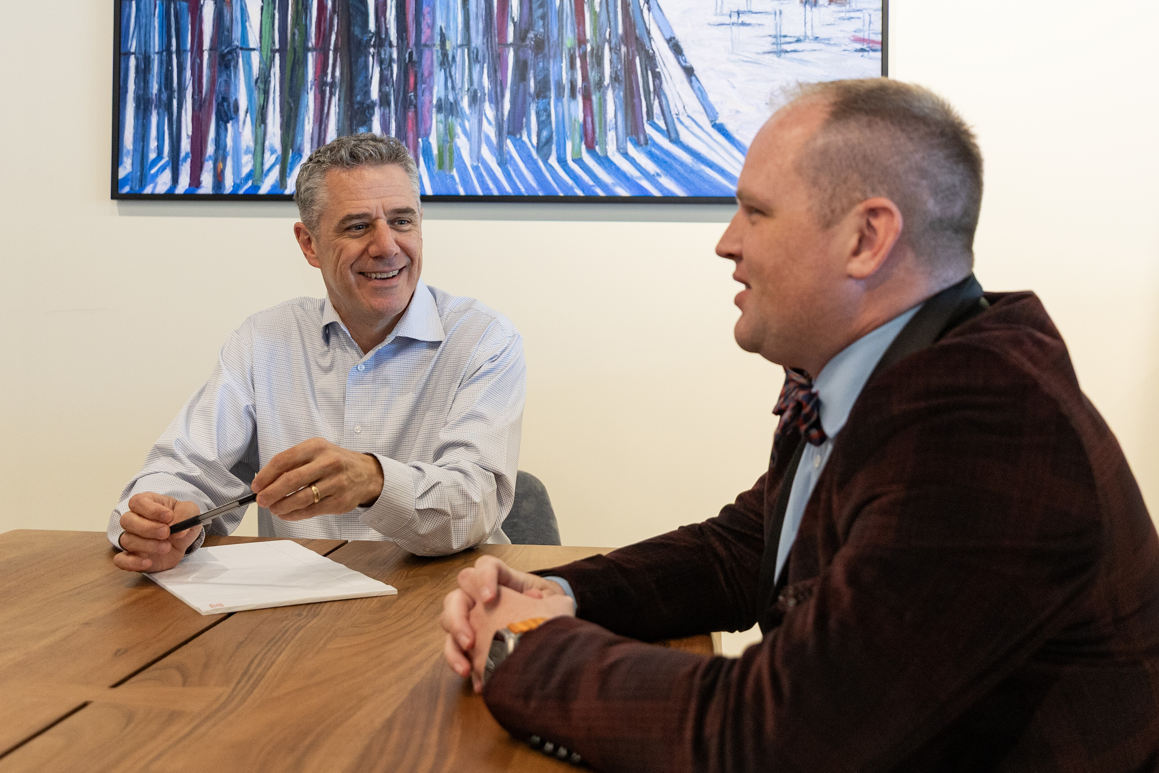 Two men sit at a wooden table, engaged in a conversation, smiling at each other.