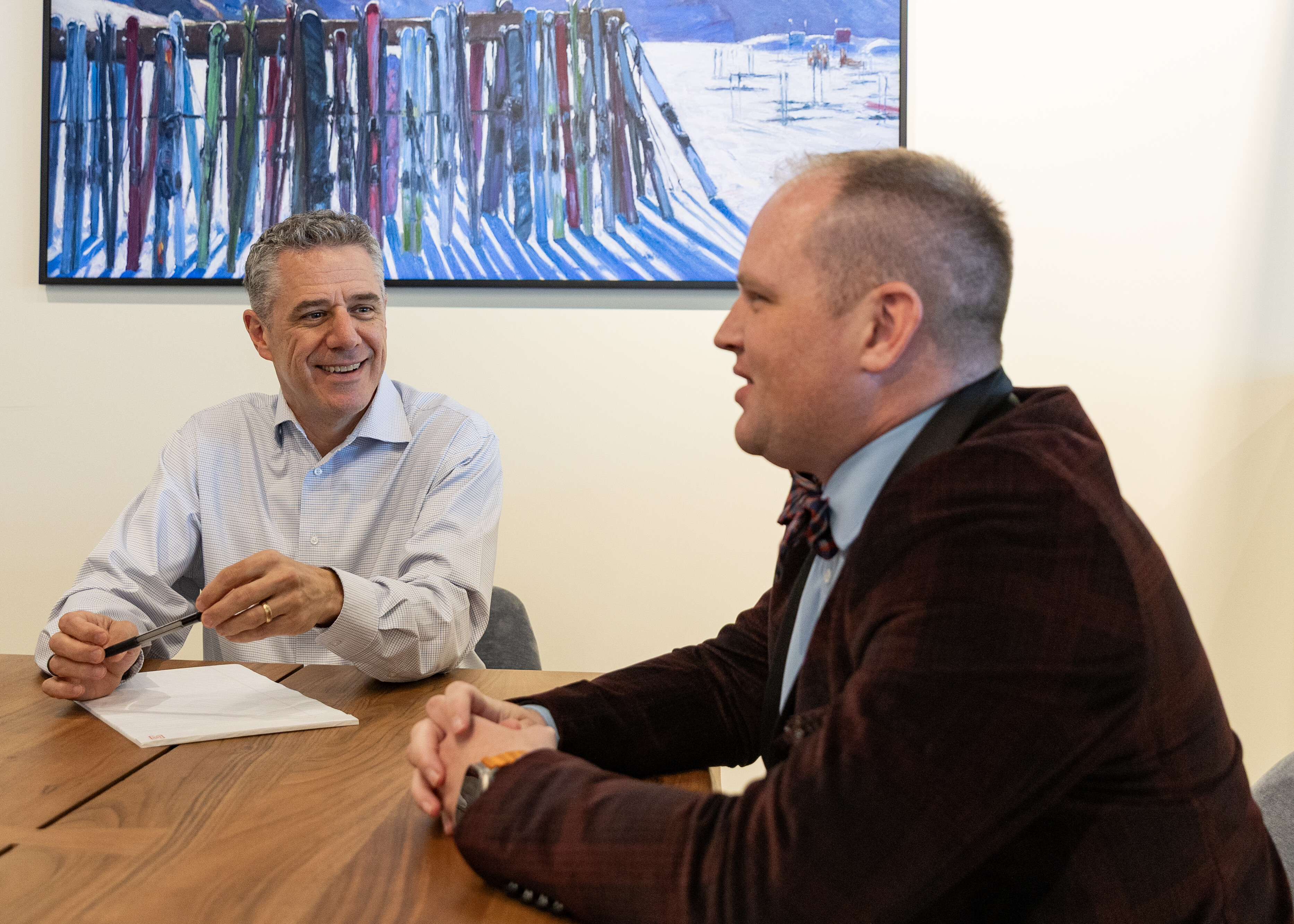 Two men in a meeting, smiling and discussing over a wooden table, with artwork in background.