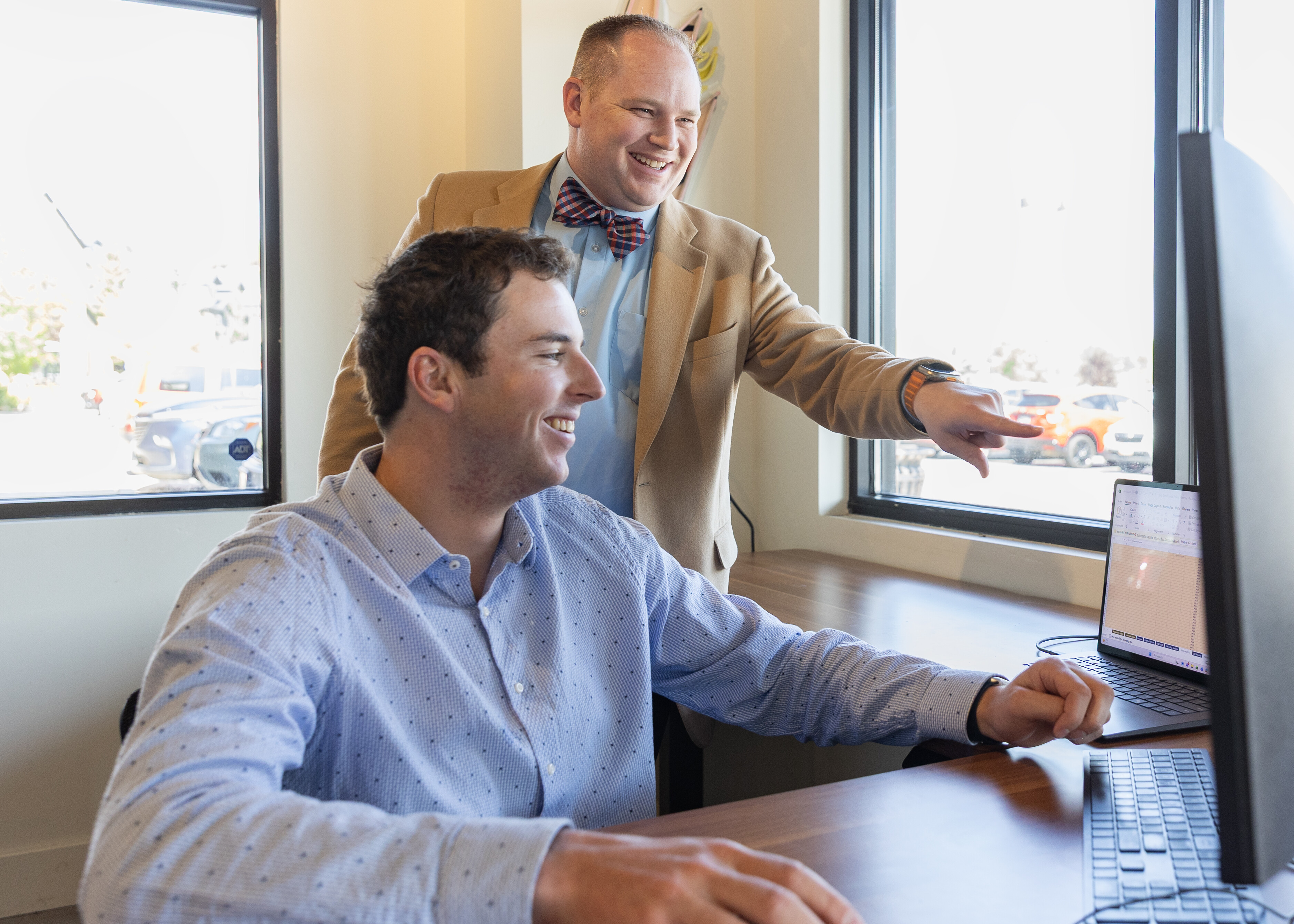 Two men collaborate at a desk, one pointing at a laptop screen while smiling.